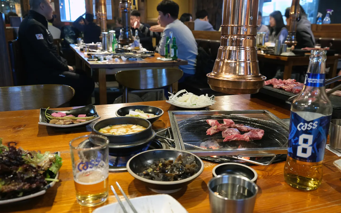 Korean barbecue table with charcoal grill, side dishes, and drinks.