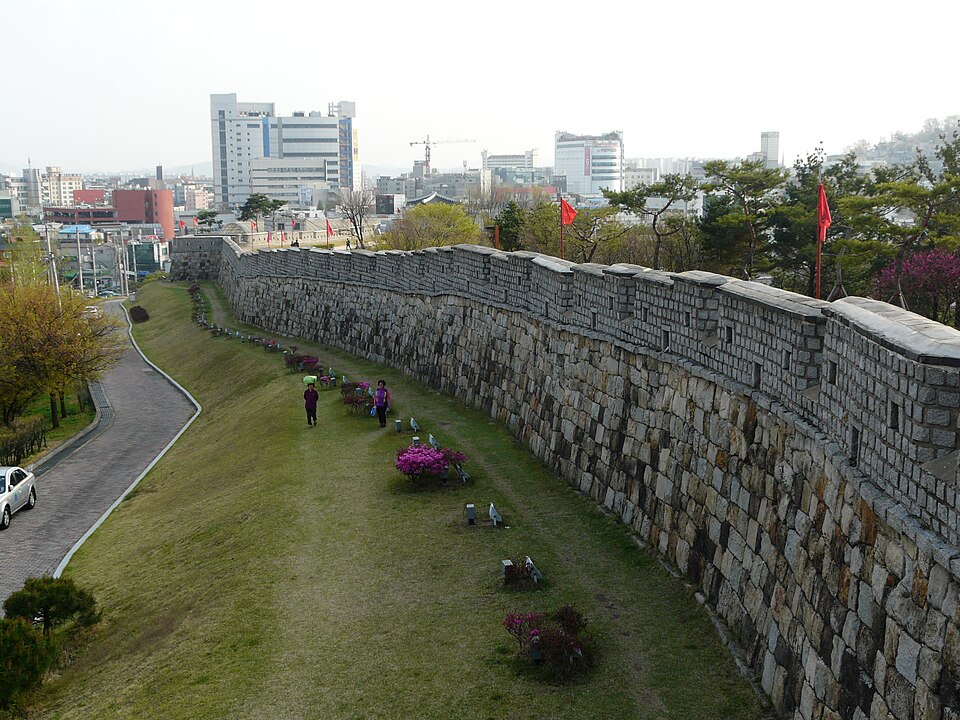 Visitors walking beside the long stone wall of Suwon Hwaseong Fortress, with the modern city visible beyond.